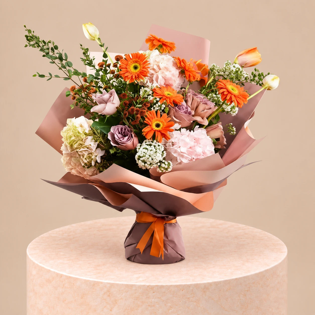 A vibrant floral bouquet featuring orange gerbera daisies, pink and white carnations, lavender-purple roses, and green foliage, wrapped in peach and brown paper with an orange ribbon, displayed on a round beige pedestal against a solid beige background. (WebP Optimized)