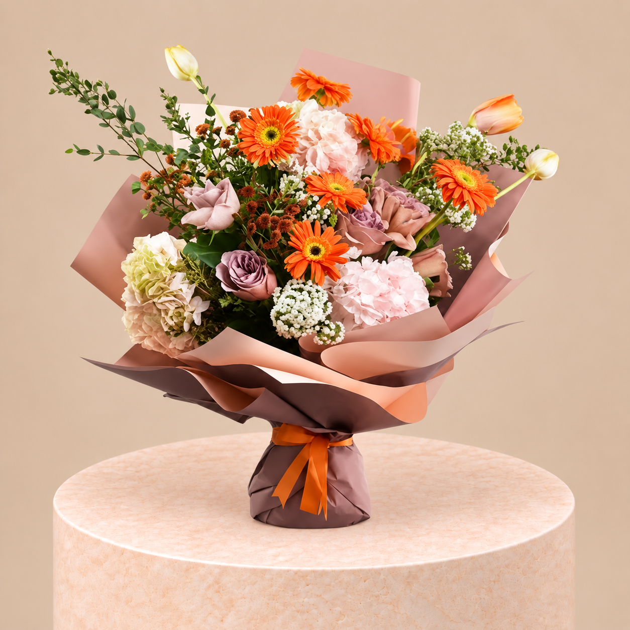 A vibrant floral bouquet featuring orange gerbera daisies, pink and white carnations, lavender-purple roses, and green foliage, wrapped in peach and brown paper with an orange ribbon, displayed on a round beige pedestal against a solid beige background.