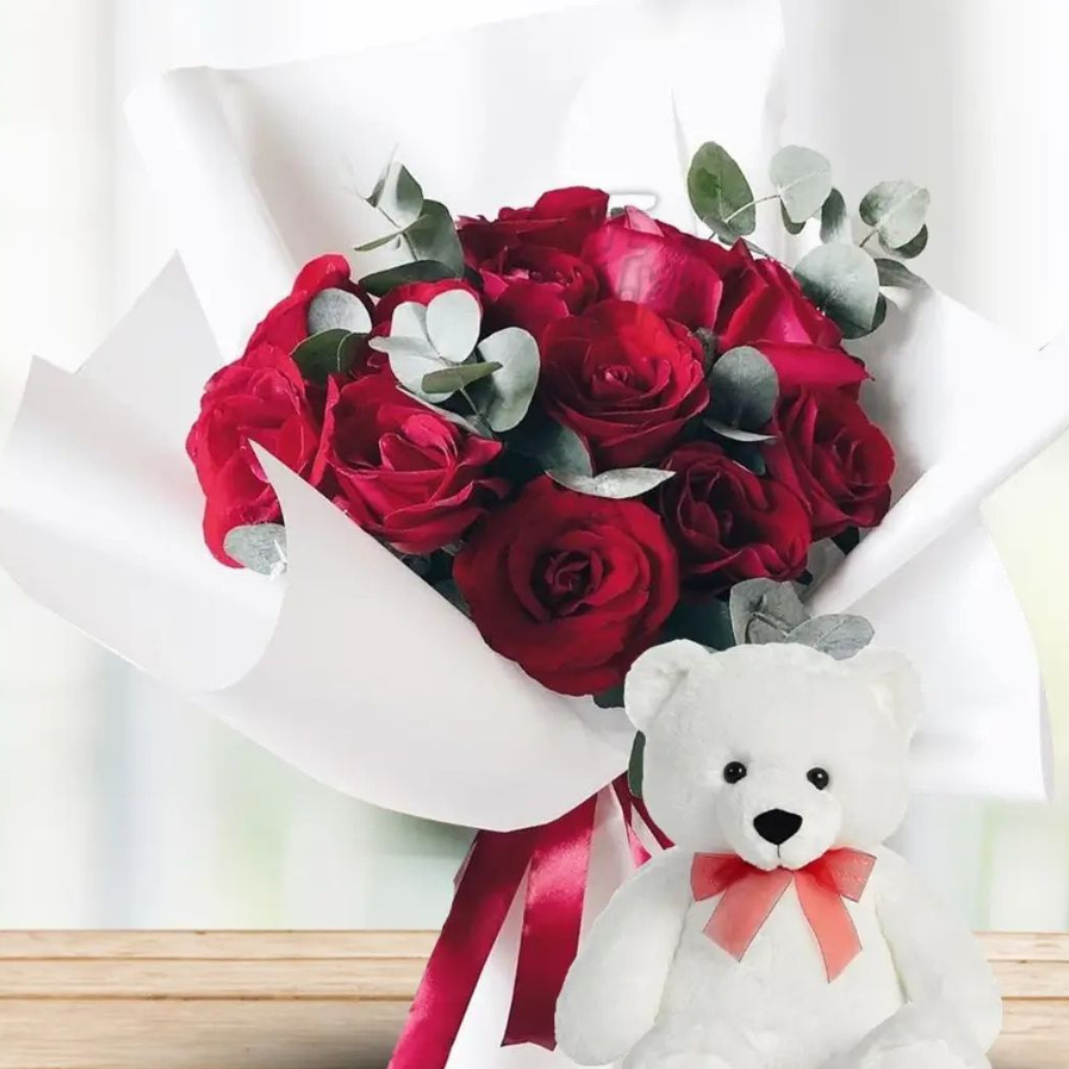 Teddy & Blooms Affair a bouquet of red roses with eucalyptus leaves and green fillers, accompanied by a white teddy bear with a red bow tie – Petal Frost flower shop