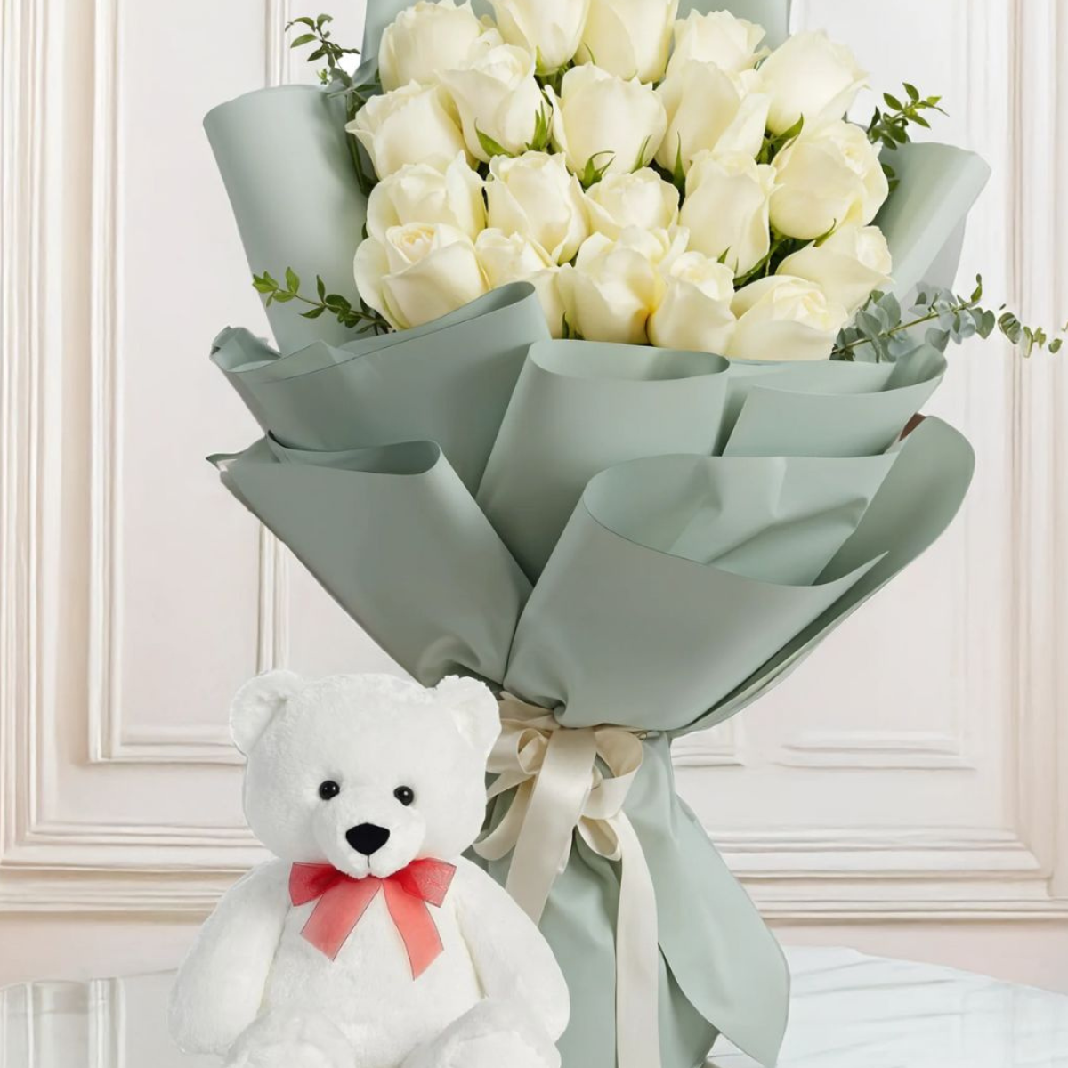 Teddy & Blooms Harmony a bouquet of white roses with green foliage wrapped in grey paper, accompanied by a white teddy bear with a red ribbon bow on its neck – Petal Frost flower shop