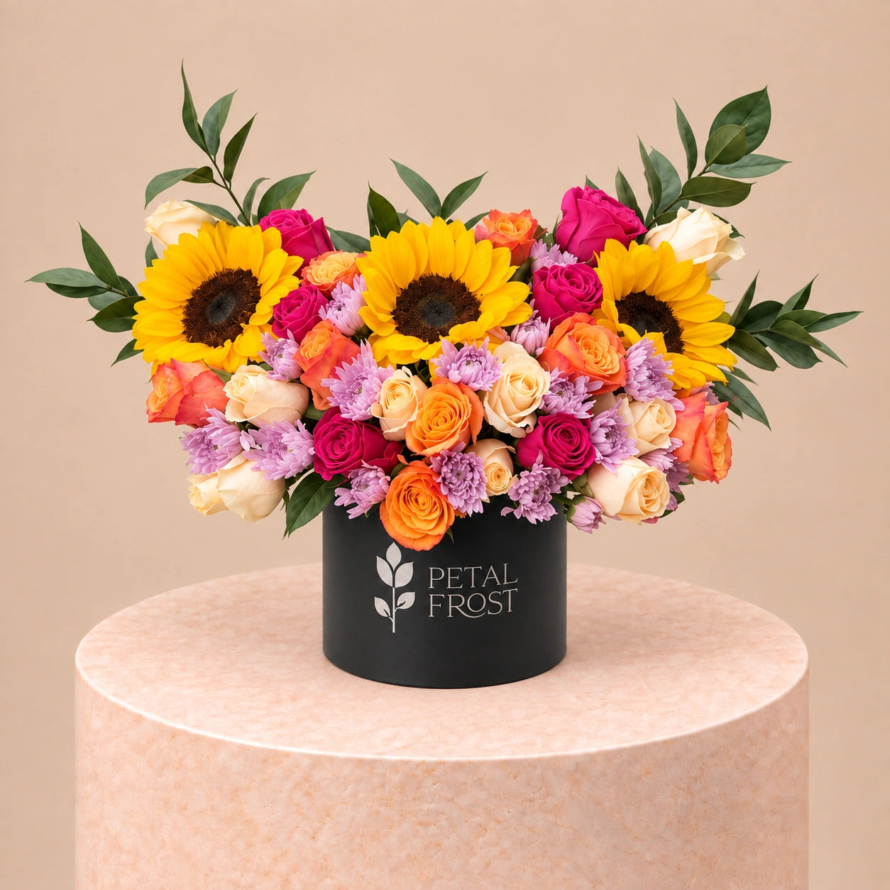 A vibrant floral arrangement featuring sunflowers, pink and orange roses, and purple filler flowers in a black cylindrical box with the brand name 'PETAL FROST' printed on the front, displayed on a round beige pedestal against a solid beige background.