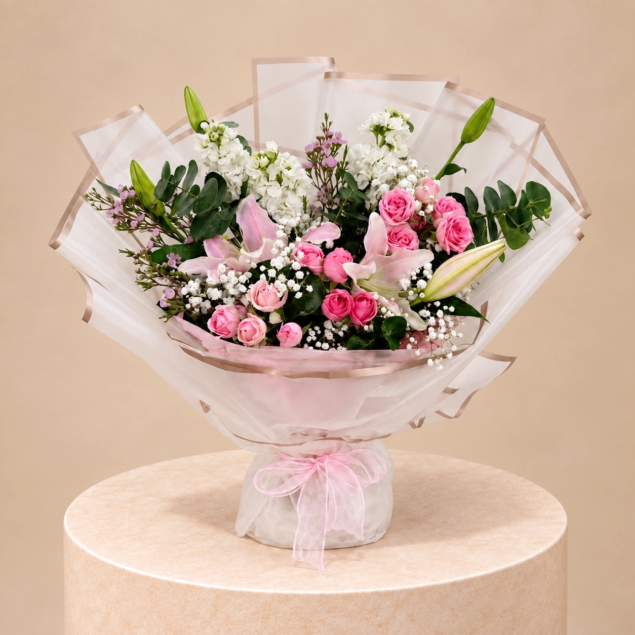 A floral bouquet wrapped in sheer white tissue paper with a pink ribbon, displayed on a round beige pedestal against a warm beige background. The bouquet features pink roses, pink lilies, white baby's breath, and green foliage.