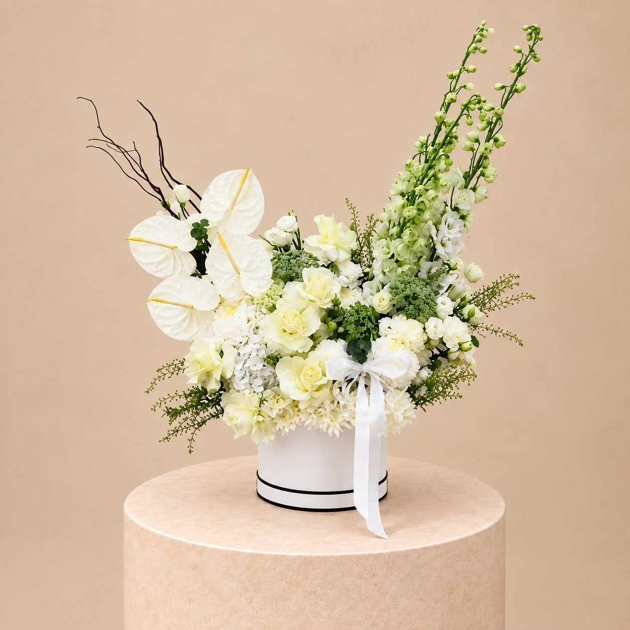A floral arrangement featuring white and pale yellow flowers with green foliage, presented in a white cylindrical box with a white ribbon bow, set on a round beige pedestal against a solid beige background.